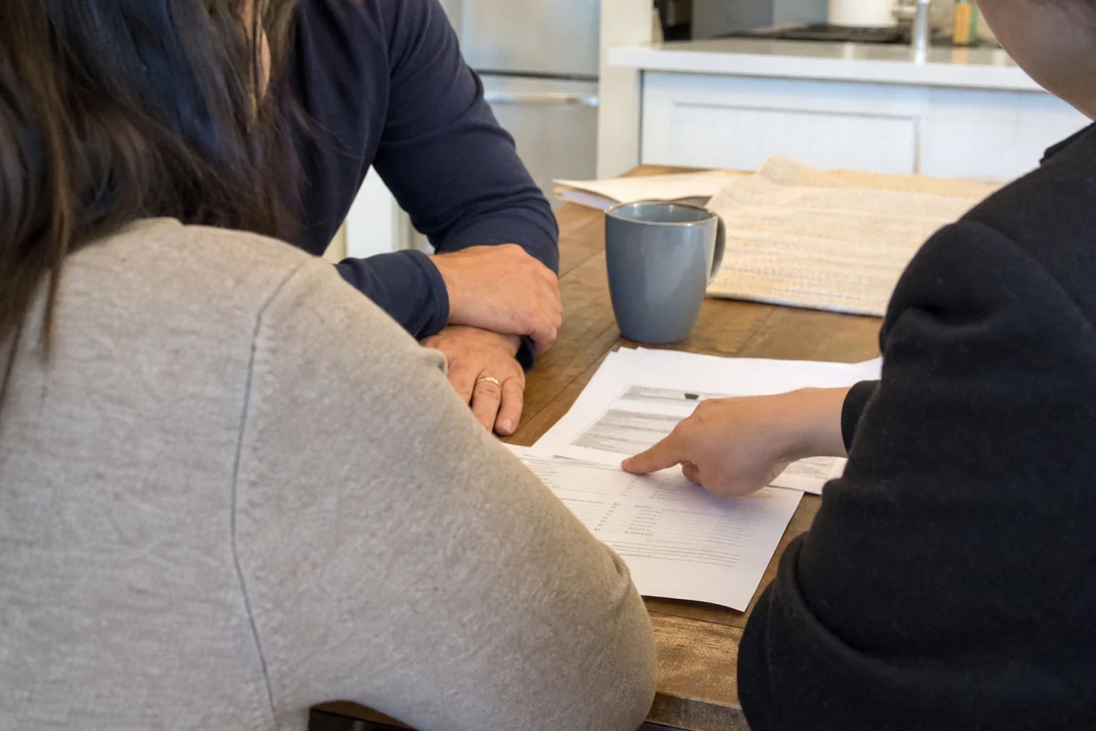 Over-the-shoulder view of a couple at a wooden kitchen table as an agent points to a line on printed papers, with a gray coffee mug nearby and a white kitchen behind