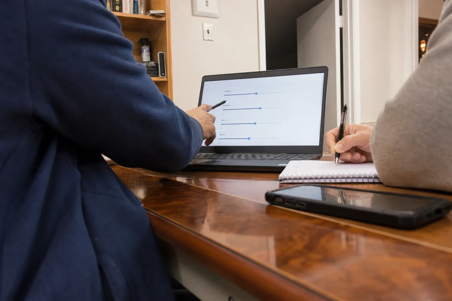 Person in a navy jacket points a pen at slider settings on a laptop while another person takes notes in a spiral notebook at a wooden office desk